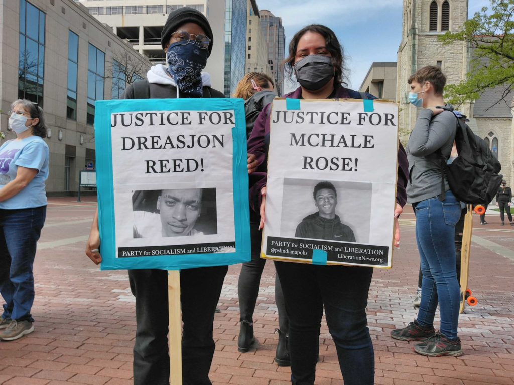 Two individuals hold banners, one reading "Justice for Dreasjon Reed" (left) and the other reading "Justice for McHale Rose" (right)
