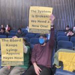 Stephen Lane holds up yellow placard at a City County Council, reading "The system is broken: We need a new one!"
