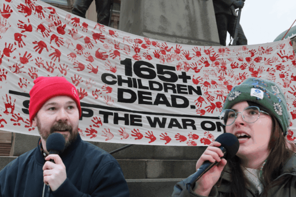 Two organizers speak in front of a banner against the war in Iran