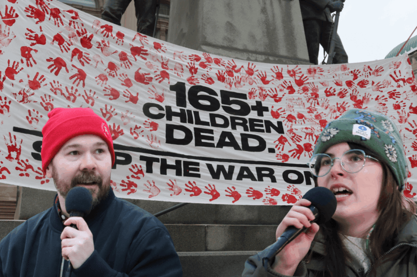 Two organizers speak in front of a banner against the war in Iran
