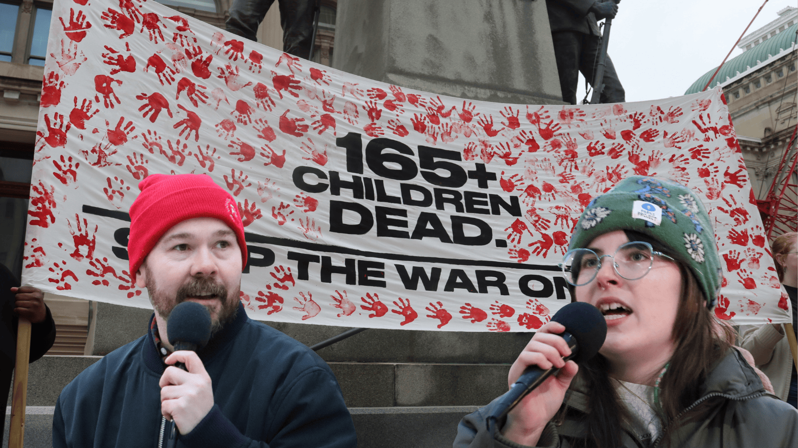 Two organizers speak in front of a banner against the war in Iran